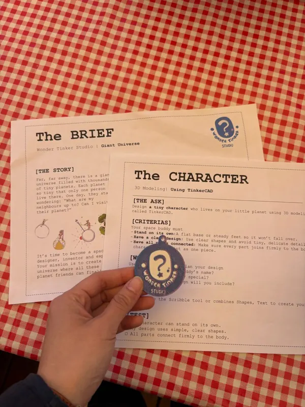 A hand holds a Wonder Tinker token over the project brief and character design sheet